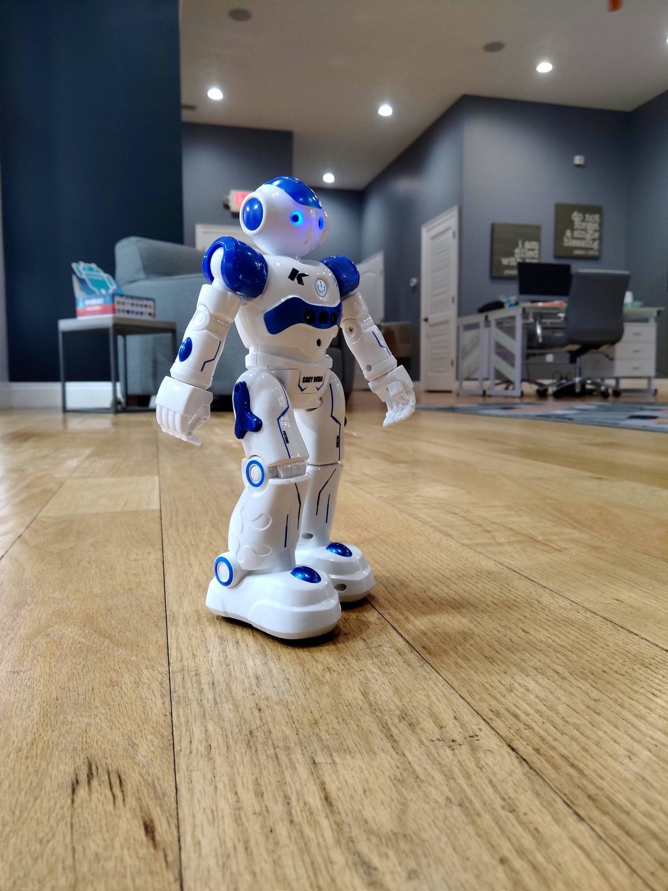 A small white and blue robot standing on the wood floor of the Mica Specialties office